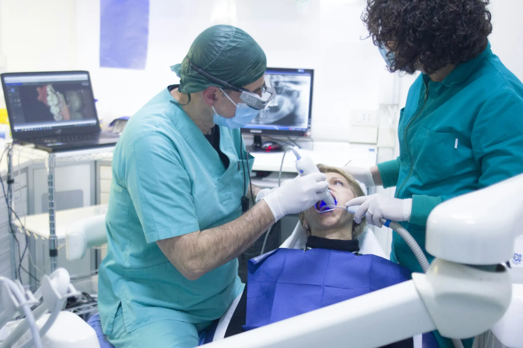 A dentist and an assistant in scrubs treating a patient in the chair, an X-ray on the screen behind them.