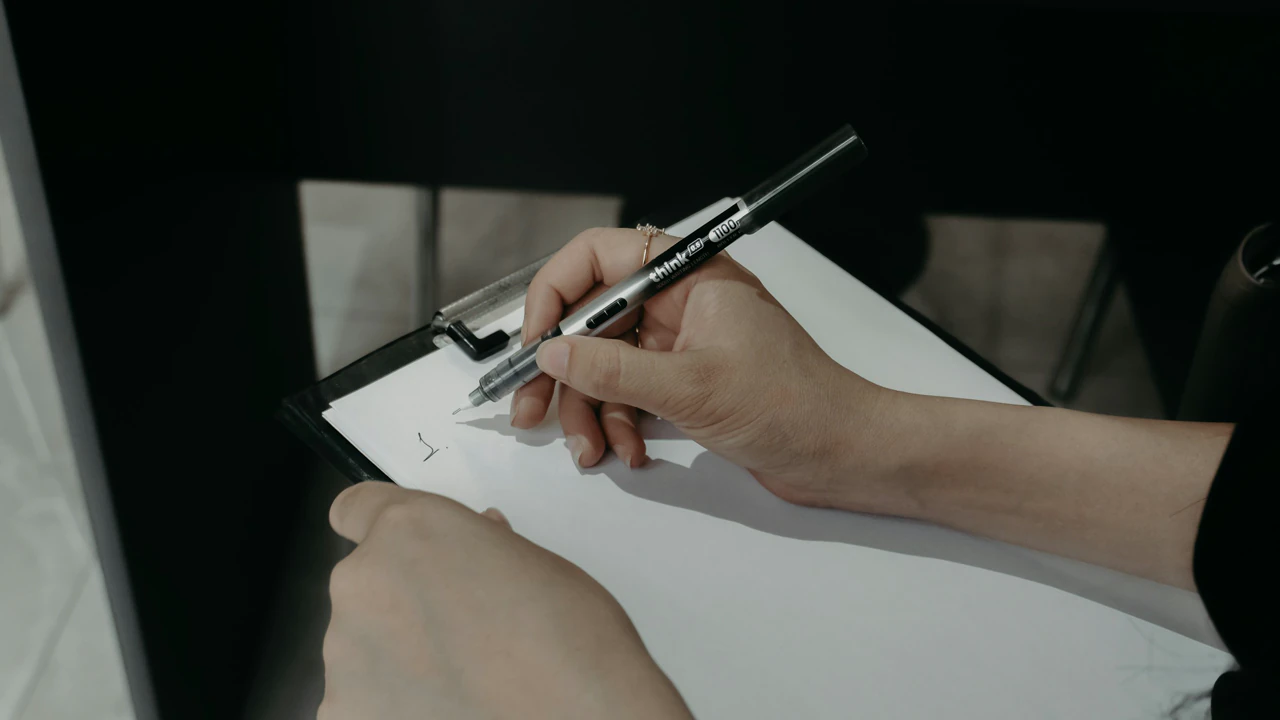 A hand writing on a clipboard with a black pen, in low light.