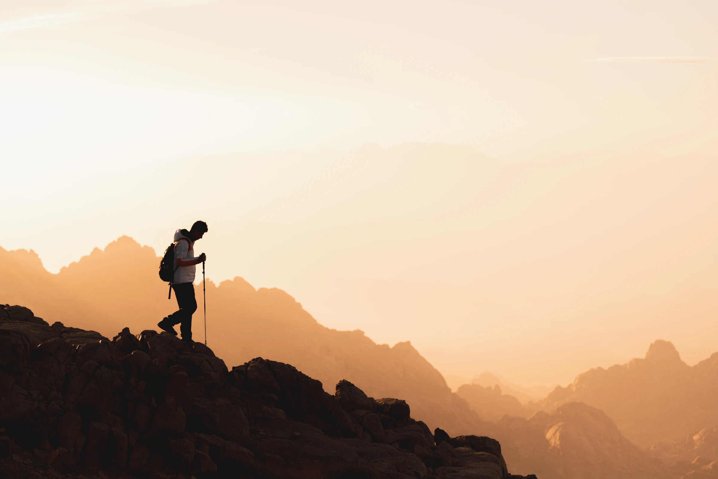 A hiker with poles and a pack climbing a ridge toward a summit at sunrise.
