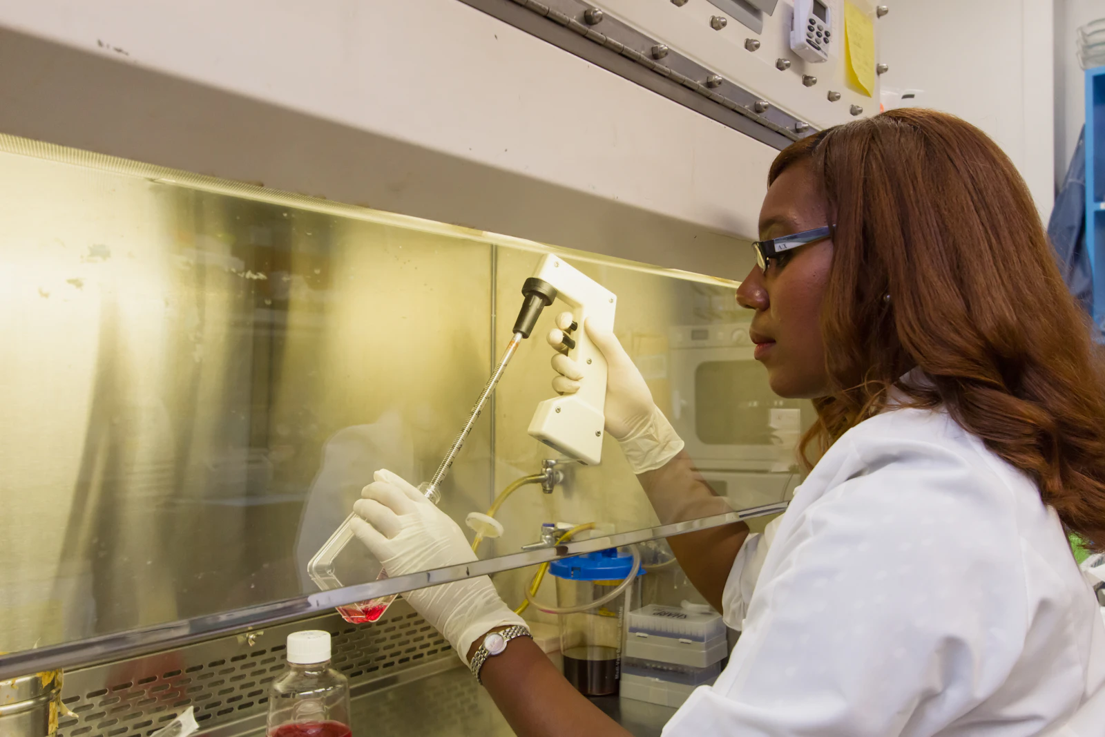 A researcher in a white coat pipetting inside a laboratory hood, seen in profile.