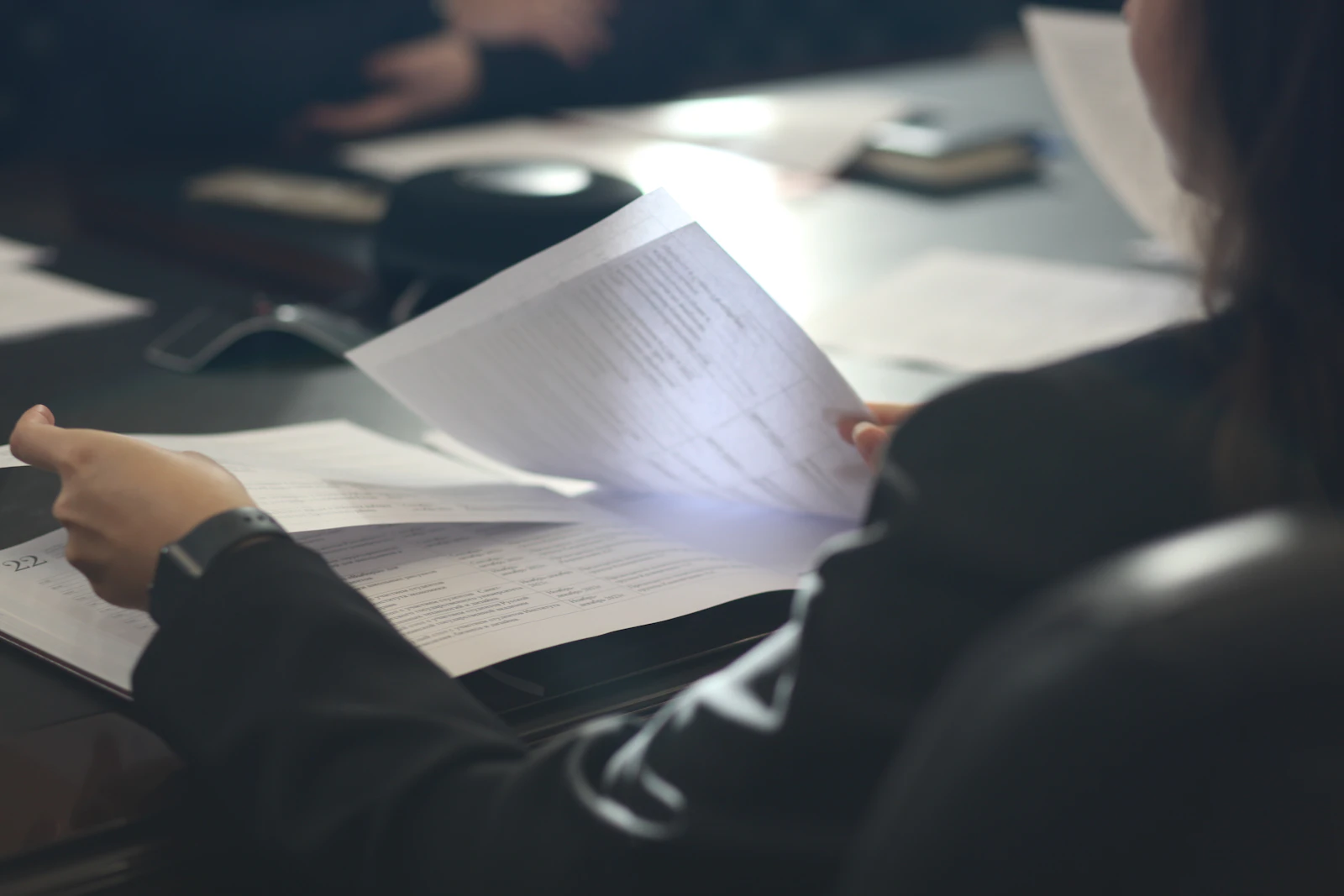A person at a meeting table turning printed pages, seen from behind the shoulder.