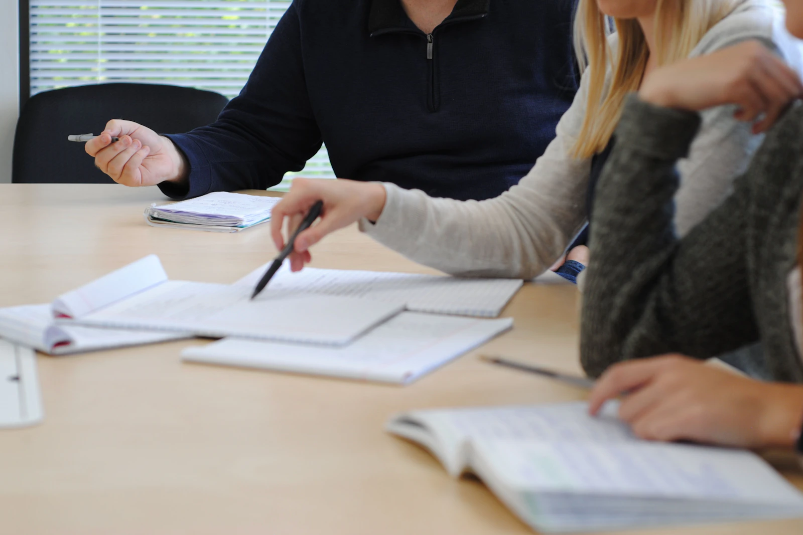 Hands at a meeting table, one pointing a pen at an open notebook among papers.