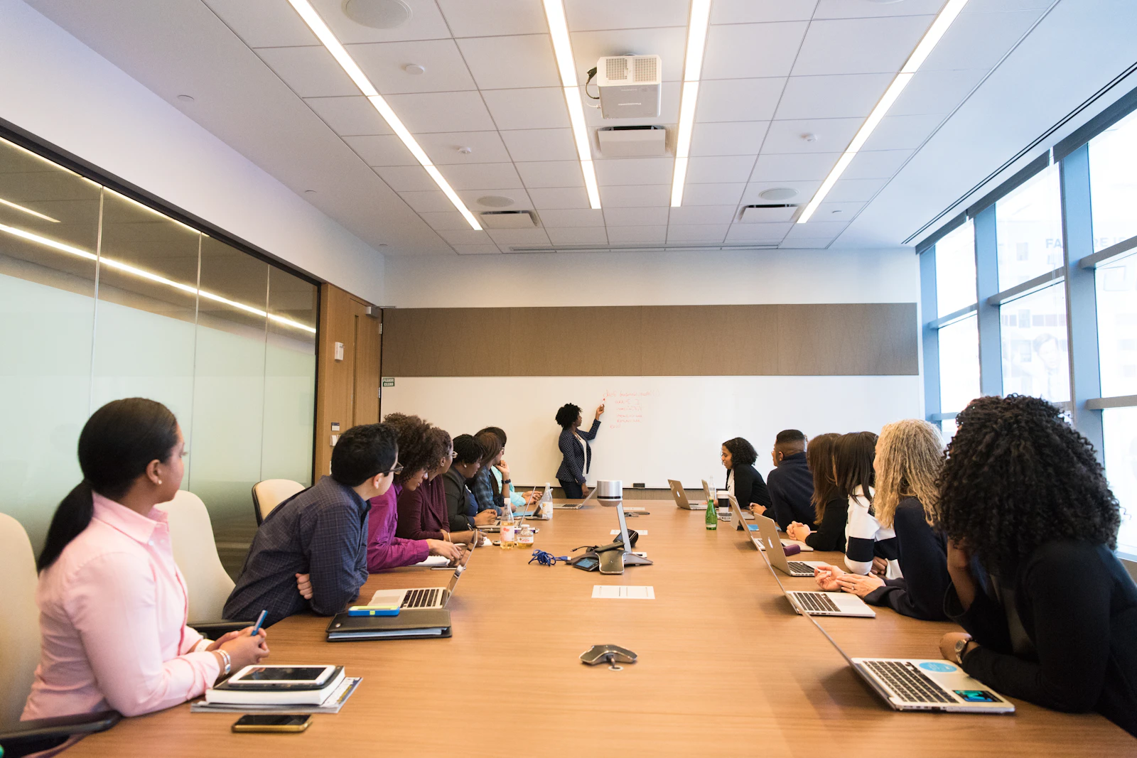 A long boardroom table seen from one end, people along both sides turned toward a colleague at the whiteboard.