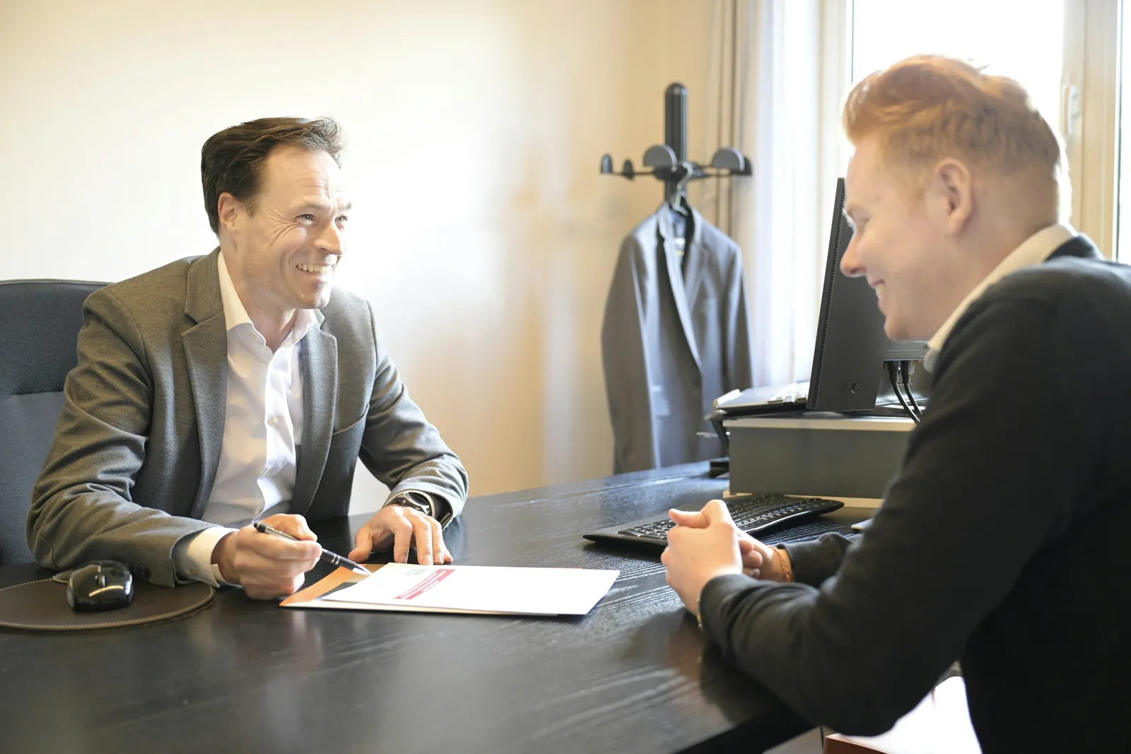 Two men in conversation across an office desk, one holding a pen over a printed page.